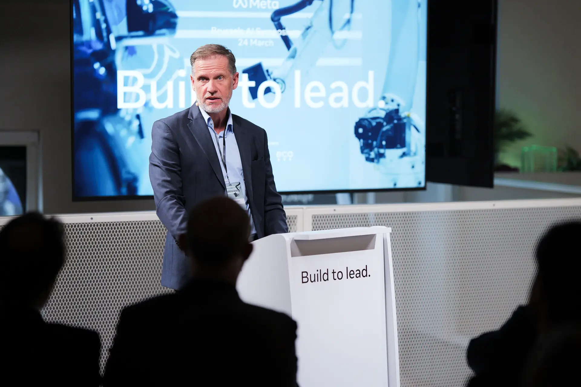 A speaker at a lectern marked &ldquo;Build to lead.&rdquo;, with the symposium title on the screen behind