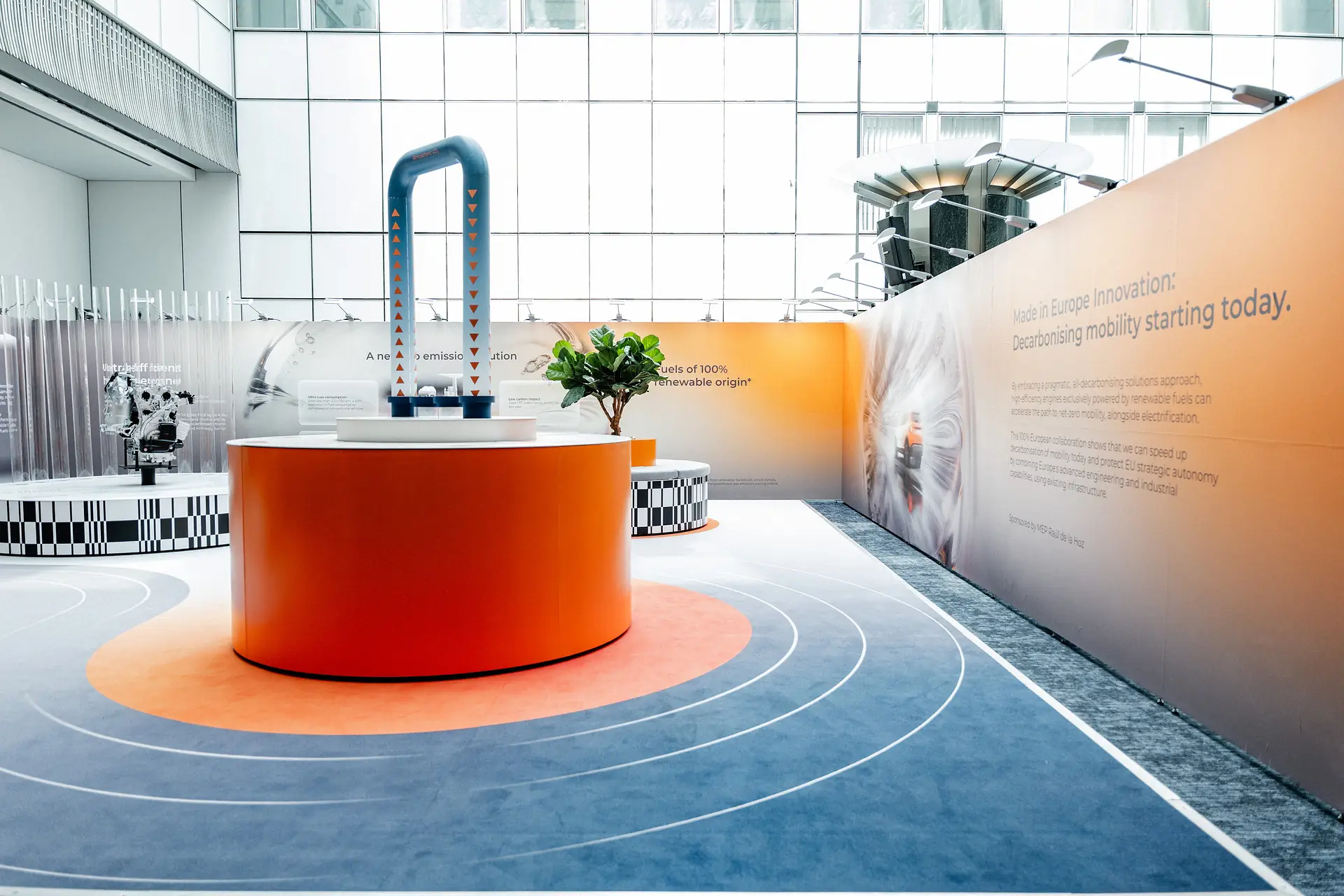 The exhibition stand in the European Parliament: an orange plinth carrying the net zero CO2 cycle sculpture, beneath a wall reading &ldquo;Made in Europe Innovation: Decarbonising mobility starting today&rdquo;