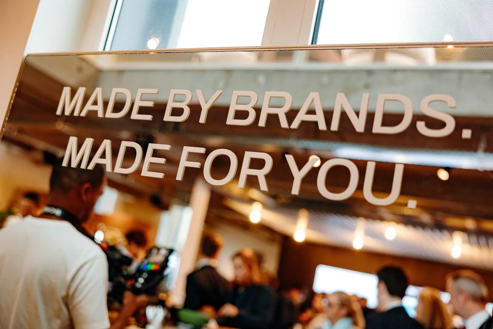 A sign reading &ldquo;Made by brands. Made for you.&rdquo;, with a camera crew filming beyond