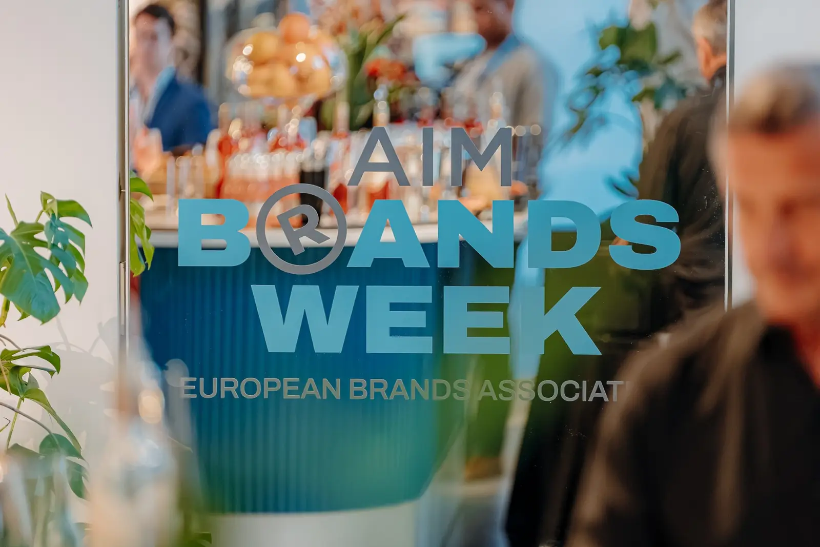 The AIM Brands Week logo on the glass, with guests behind