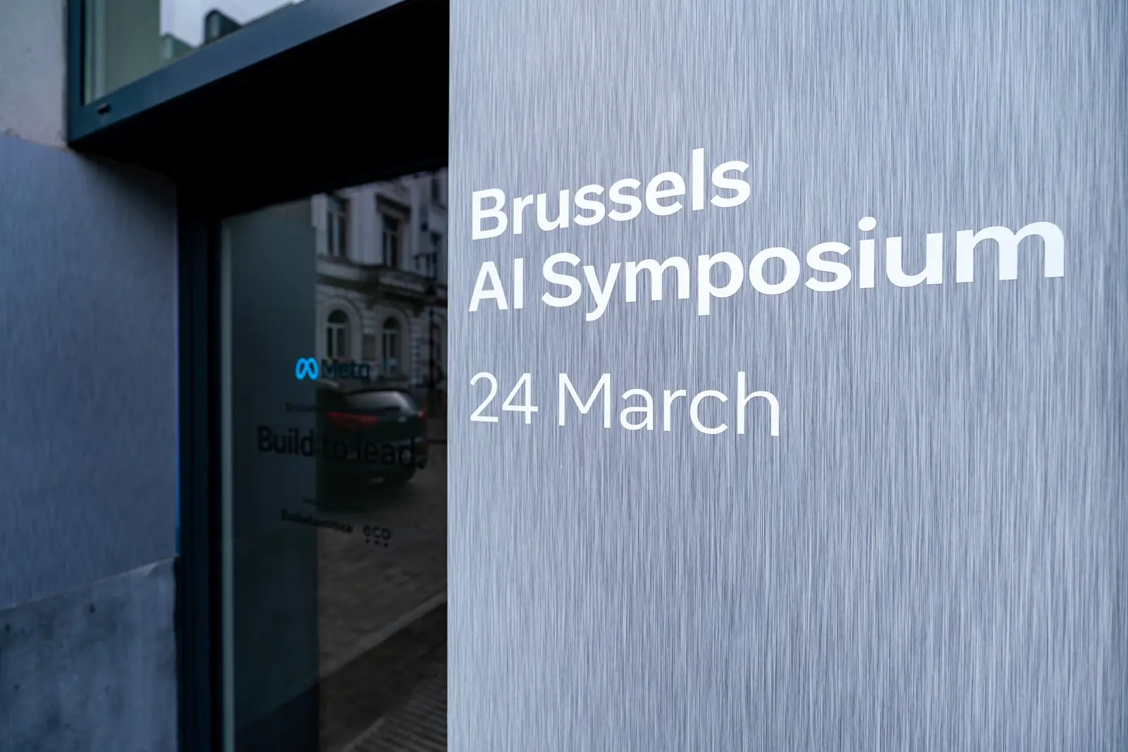 A brushed metal sign reading &ldquo;Brussels AI Symposium, 24 March&rdquo;