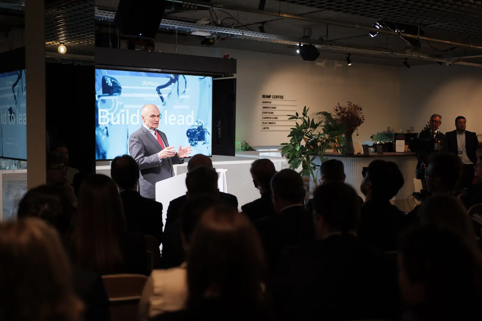 A speaker addressing a full room, with &ldquo;Build to lead.&rdquo; on the screen behind