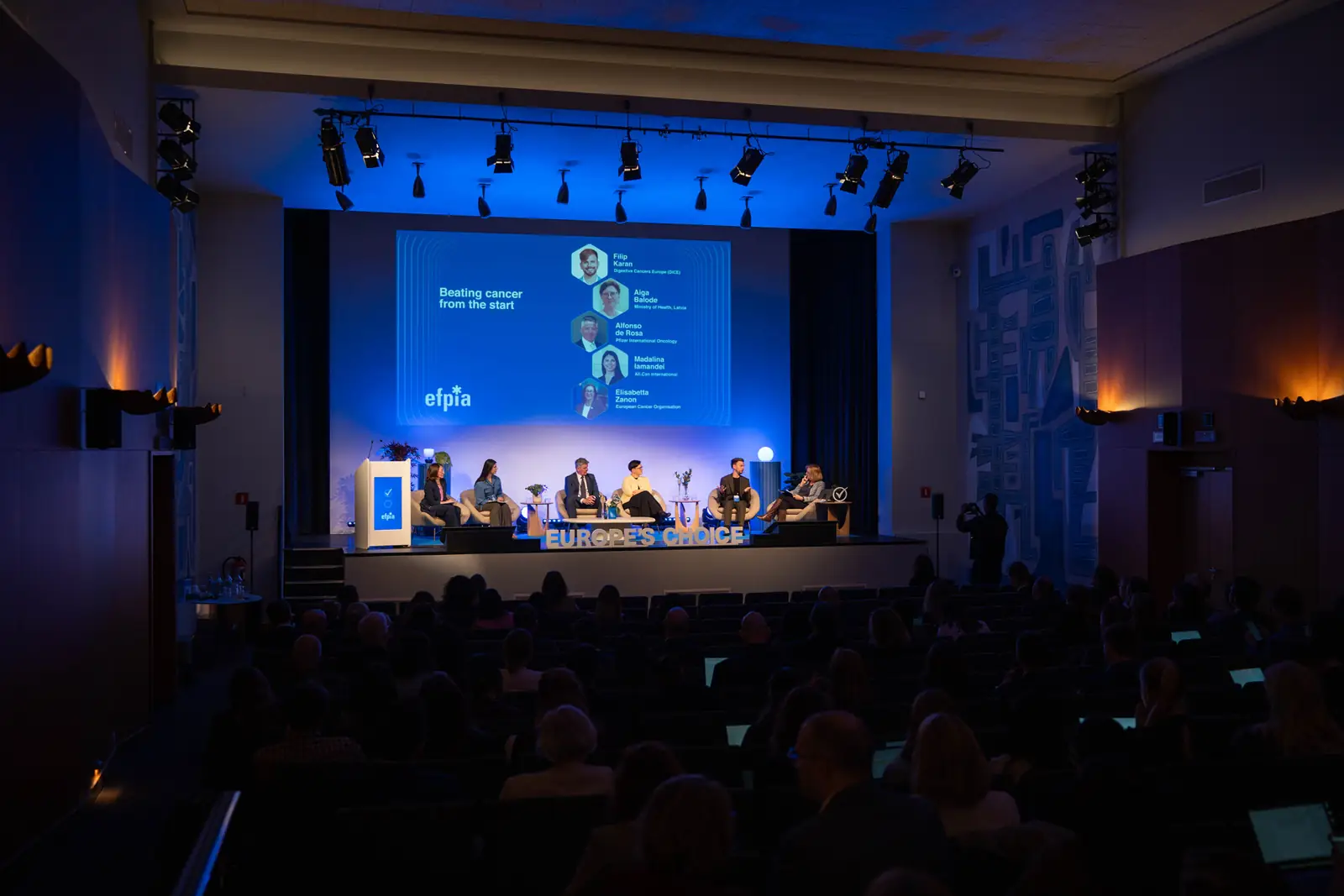 The main stage in a packed auditorium, with &ldquo;Beating cancer from the start&rdquo; on the screen
