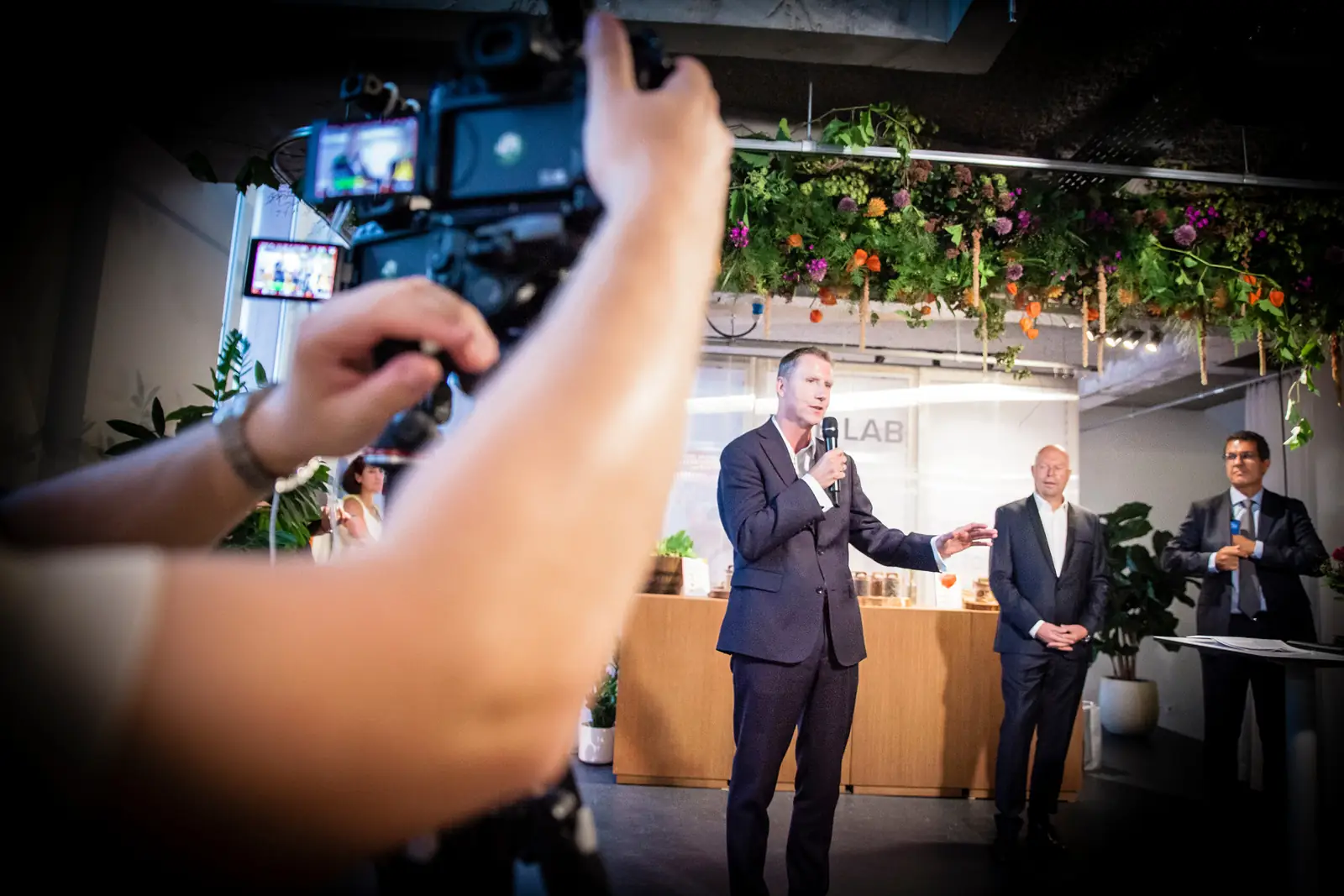A speaker addressing the room under a canopy of dried flowers, filmed from the crowd